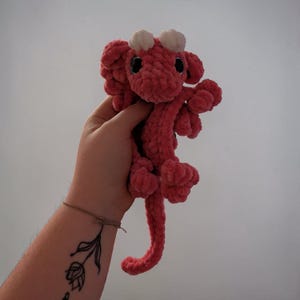 May include: A small, handmade crochet dragon plush toy crafted from soft, textured pink yarn. The creature features two small white horns on its head, large black safety eyes, and a long, curled tail. The design includes detailed wings and limbs.