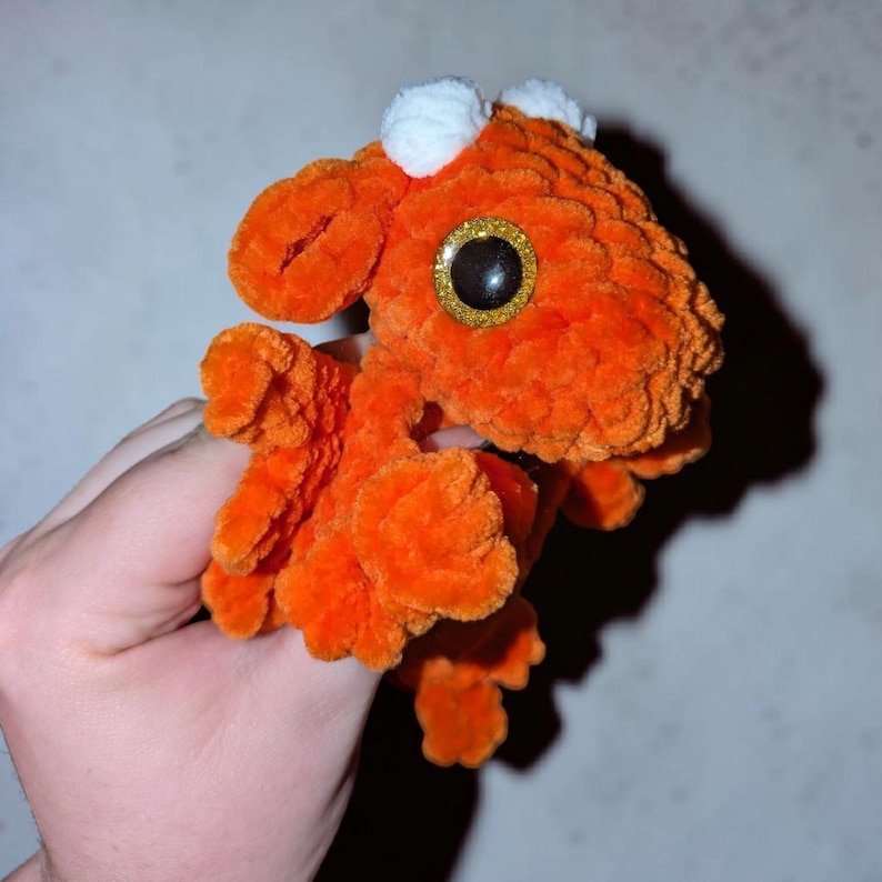 May include: A small, bright orange crocheted plush toy shaped like a dragon. The creature features a textured, fuzzy yarn surface, two small white tufts on its head, and a single large, round eye with a black pupil and a gold glittery iris.