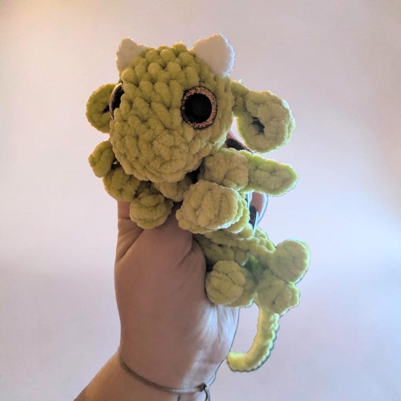May include: A light green crocheted plush dragon toy featuring a soft, textured yarn finish, two small white horns on the top of the head, and large, round, dark brown eyes with a subtle glittery rim, crafted in a whimsical, rounded creature shape.