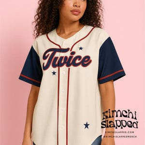 May include: Cream-colored baseball jersey with navy blue sleeves and red accents. The word "Twice" is written across the chest in a stylized font, with star graphics. The jersey has a button-up front and the text "kimchi slapped" is visible.