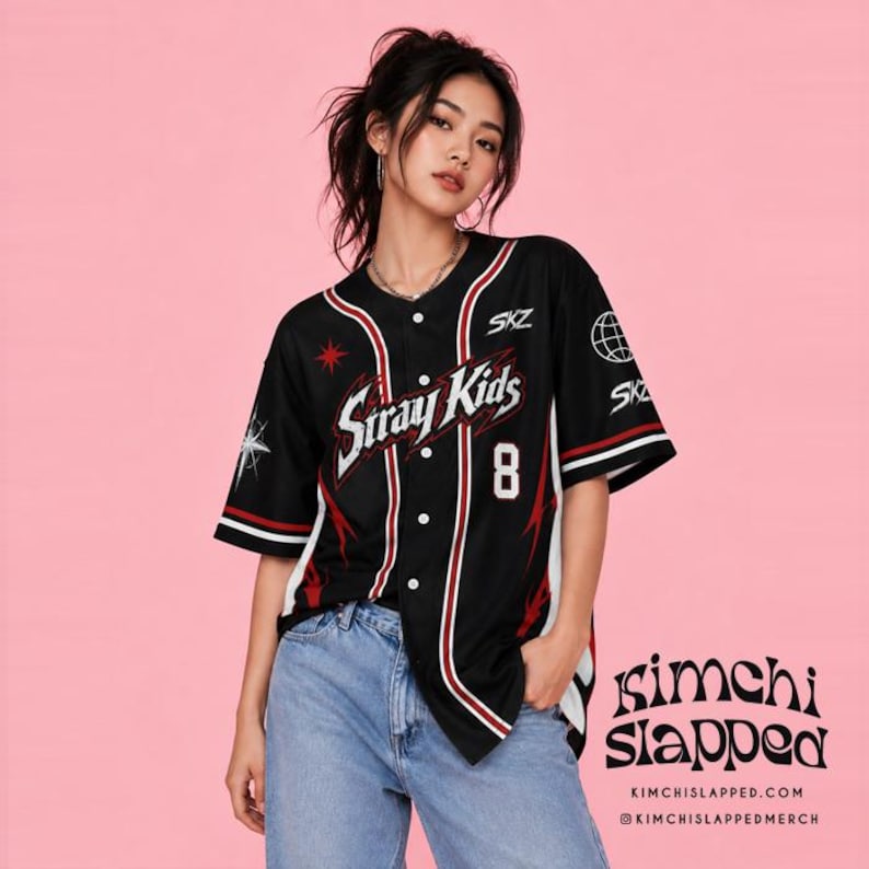 May include: A black baseball jersey featuring white and red vertical stripes, the text Stray Kids, the number 8, and star graphics. The sleeves display a globe icon and the text SKZ. The shirt has a button-down front and is paired with blue jeans.