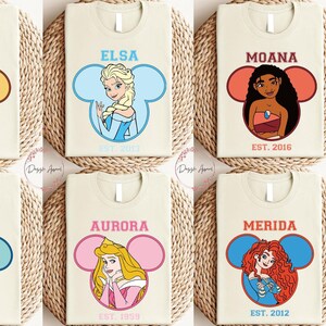 May include: Six cream-colored t-shirts with a Disney princess character in a Mickey Mouse ear silhouette. Each shirt has the princess's name and the year "EST." followed by a year.