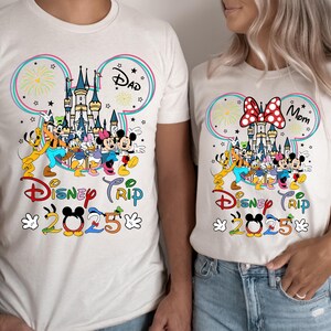 Op de afbeelding: Witte T-shirts met een Mickey Mouse-oorontwerp met een kleurrijke illustratie van Assepoester's kasteel, vuurwerk en Mickey Mouse-personages. De tekst "Disney Trip 2025" staat op de shirts gedrukt. E&eacute;n shirt zegt "Papa" en het andere zegt "Mama".