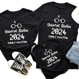 May include: Black t-shirts with white text that reads "Universal Studios 2024 FAMILY VACATION". The design includes glasses and a lightning bolt. The image also shows a white hat, blue jeans, and black sneakers.