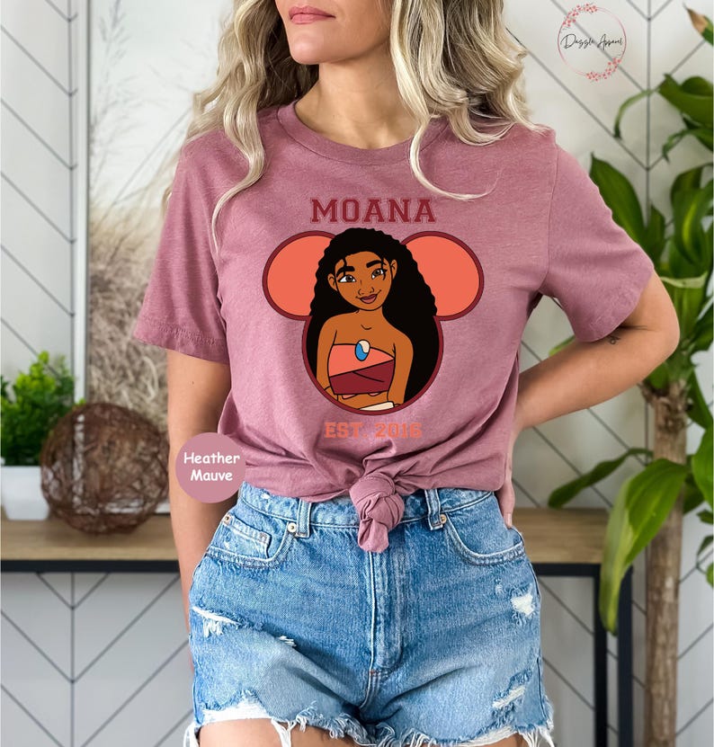 May include: A heather mauve t-shirt with a graphic of Moana from the Disney movie Moana. The graphic is in a Mickey Mouse ear shape with the text "Moana Est. 2016".