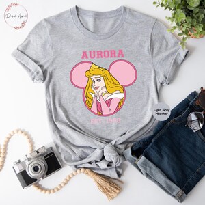 May include: A light gray heather t-shirt with a pink and white Mickey Mouse ear design featuring a cartoon image of Princess Aurora from Sleeping Beauty. The text "AURORA EST. 1959" is printed on the shirt.