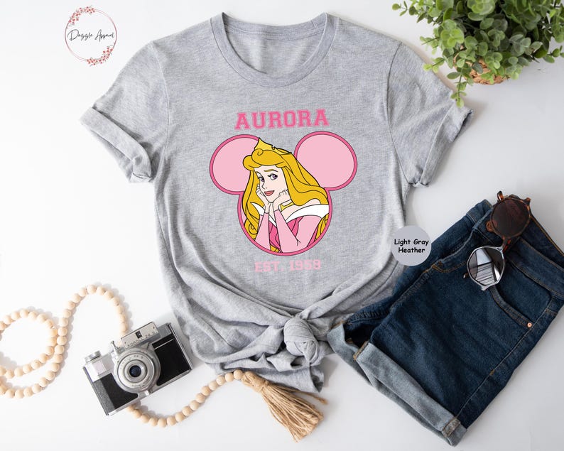 May include: A light gray heather t-shirt with a pink and white Mickey Mouse ear design featuring a cartoon image of Princess Aurora from Sleeping Beauty. The text "AURORA EST. 1959" is printed on the shirt.