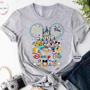Op de afbeelding: Lichtgrijs heather t-shirt met een Mickey Mouse-oorsilhouet, een kleurrijke illustratie van Assepoester's kasteel, Mickey Mouse en andere Disney-personages, en de tekst "Disney Trip 2025".