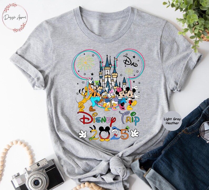Op de afbeelding: Lichtgrijs heather t-shirt met een Mickey Mouse-oorsilhouet, een kleurrijke illustratie van Assepoester's kasteel, Mickey Mouse en andere Disney-personages, en de tekst "Disney Trip 2025".
