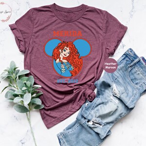 May include: Heather maroon t-shirt with a graphic of Merida from Disney's Brave in a Mickey Mouse ear silhouette. The text "Merida" and "est. 2023" are printed on the shirt.