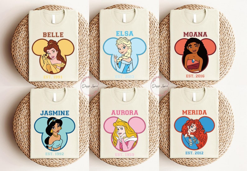 May include: Six cream-colored t-shirts with a Disney princess character in a Mickey Mouse ear silhouette. Each shirt has the princess's name and the year "EST." followed by a year.