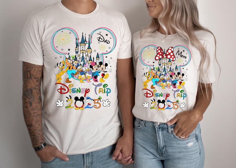 Op de afbeelding: Witte T-shirts met een Mickey Mouse-oorontwerp met een kleurrijke illustratie van Assepoester's kasteel, vuurwerk en Mickey Mouse-personages. De tekst "Disney Trip 2025" staat op de shirts gedrukt. E&eacute;n shirt zegt "Papa" en het andere zegt "Mama".