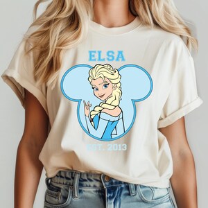 May include: A white t-shirt with a blue Mickey Mouse ear silhouette. Inside the ear is a cartoon image of Elsa from Frozen. The text "Elsa" is above the image and "Est. 2013" is below the image.