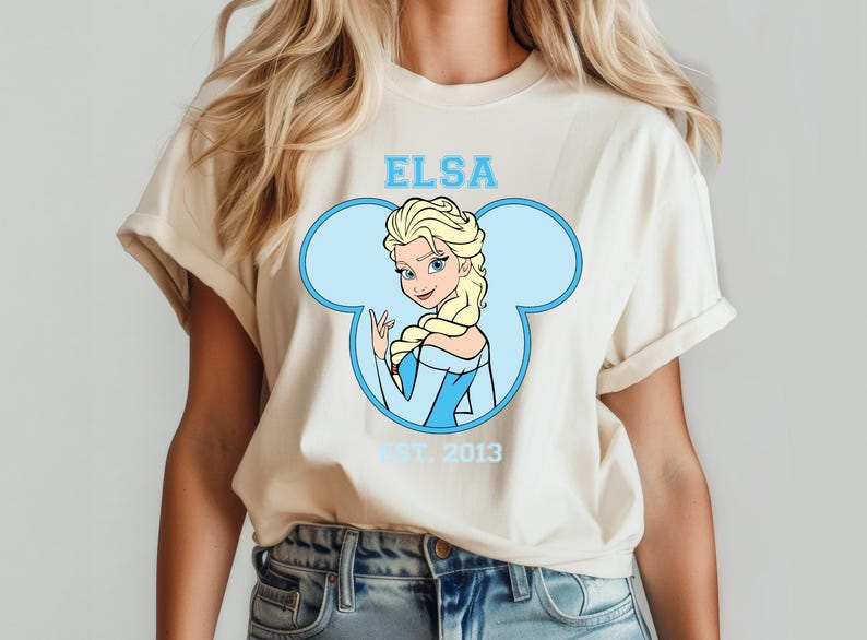 May include: A white t-shirt with a blue Mickey Mouse ear silhouette. Inside the ear is a cartoon image of Elsa from Frozen. The text "Elsa" is above the image and "Est. 2013" is below the image.