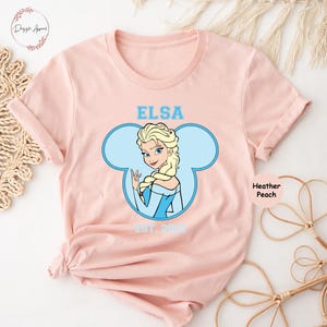 May include: A pink t-shirt with a blue Mickey Mouse head design featuring Elsa from Frozen. The text "Elsa Est. 2013" is printed on the shirt.