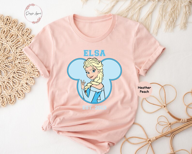 May include: A pink t-shirt with a blue Mickey Mouse head design featuring Elsa from Frozen. The text "Elsa Est. 2013" is printed on the shirt.