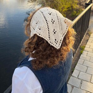 Op de afbeelding: Crèmekleurige, handgebreide hoofddoek met een geometrisch openwerk ontwerp. De driehoekige sjaal wordt op het hoofd gedragen, met de punt aan de achterkant. De hoofddoek is gemaakt van zacht, getextureerd garen.