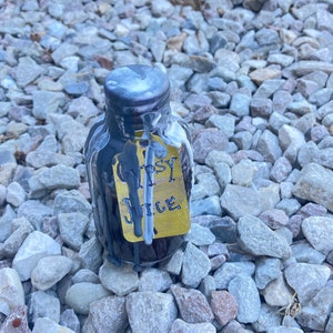 May include: A small, clear glass bottle with a silver cap and black liquid inside. The bottle has a yellow tag that reads "Gypsy Juice" in black lettering. The bottle is surrounded by small, gray rocks.