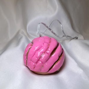 Könnte beinhalten: Ein rosa Concha-Ornament mit silbernem Band zum Aufhängen. Das Ornament hat eine strukturierte, geriffelte Oberfläche mit braunen Akzenten, die an ein traditionelles mexikanisches Süßbrot erinnert. Auf weißem, seidigem Hintergrund.
