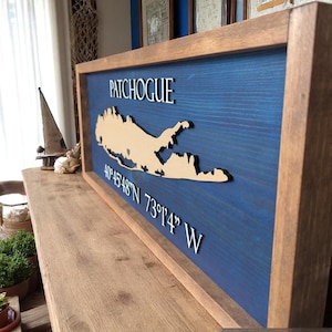 May include: Wooden framed sign featuring the word "Patchogue" in white lettering, with a beige-colored map of the area on a blue background. Coordinates are also displayed. The frame is a dark brown color.