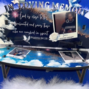 May include: A wooden memorial bench with a blue and white sky background. The bench has two photo frames with pictures of people and the text "IN LOVING MEMORY" at the top. The text "The Lord is close to the brokenhearted and saves those who are crushed in spirit. Psalms 34:18" is printed on the bench.