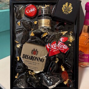 May include: A framed resin art piece featuring a miniature Disaronno bottle, Coca-Cola can pieces, and a Coke bottle cap. The artwork includes gold leaf accents and black resin, with a small black box with a gold emblem.