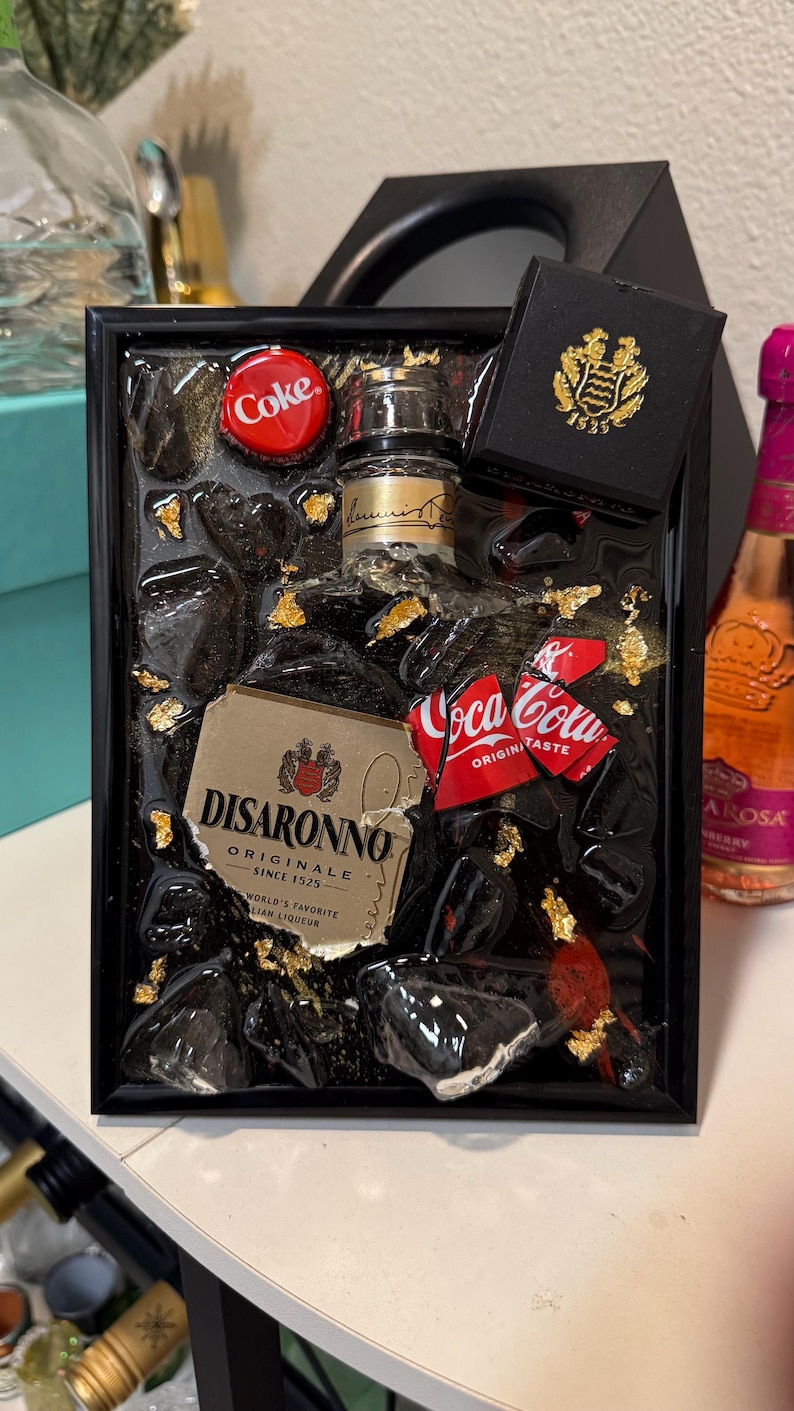 May include: A framed resin art piece featuring a miniature Disaronno bottle, Coca-Cola can pieces, and a Coke bottle cap. The artwork includes gold leaf accents and black resin, with a small black box with a gold emblem.
