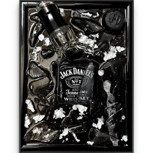 May include: Black framed artwork featuring a Jack Daniel's whiskey bottle, partially submerged in a dark liquid with silver accents. The bottle's label is prominently displayed, with the brand name and "Tennessee Sour Mash Whiskey" text.