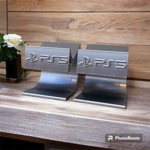 Könnte beinhalten: Zwei silberfarbene PlayStation 5 Konsolenständer mit dem PlayStation-Logo auf der Vorderseite. Die Ständer stehen auf einer hellbraunen Holzoberfläche.