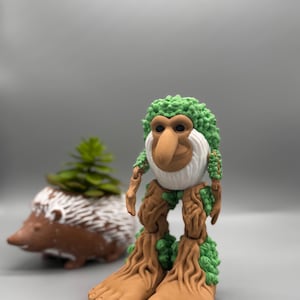 Pode incluir: Uma figura fantasiosa com um nariz grande, cabelo verde e frondoso e pernas e braços semelhantes a árvores. A figura tem uma barba branca. Um vaso de ouriço-cacheiro castanho com uma planta verde está no fundo.