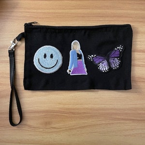 May include: Black canvas clutch with a wrist strap and zipper closure. The clutch features three embroidered patches: a light blue smiley face, a figure in a blue jacket and purple skirt, and a purple butterfly.