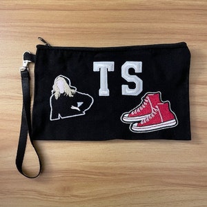 May include: Black canvas clutch with a zipper closure and wrist strap. The clutch features embroidered patches of the letters "TS", a red high-top sneaker, and a silhouette of a person.