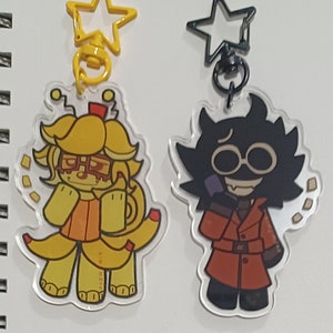 May include: Two acrylic keychains with cartoon characters. One is yellow with a banana theme, the other is black and red. Both have star-shaped clasps. The banana character has glasses and a red bow.