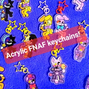 May include: A collection of colorful acrylic keychains featuring characters from the popular video game franchise Five Nights at Freddy's (FNAF). The keychains are attached to silver star-shaped charms. The text "Acrylic FNAF keychains!" is written across the image.