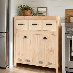 May include: A light-colored wooden cabinet with three drawers and two doors. The cabinet has black handles and hinges. A potted plant and framed picture sit on top of the cabinet. The cabinet is in a kitchen setting.