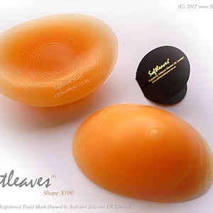 Puede incluir: Dos prótesis mamarias de silicona naranja, una con círculos concéntricos y la otra lisa. También hay un pequeño objeto negro con texto dorado. El logotipo de Softleaves y Shape X100 son visibles. También está presente el texto "Softleaves is a Registered Traid Mark Owned by Softcare Silicone UK Limited".