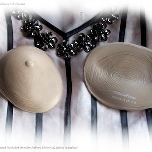 May include: Two beige silicone breast forms with a textured surface. The forms are on a white and black striped fabric. The text "Softcare Shape UK12 67110" is printed on one of the forms.