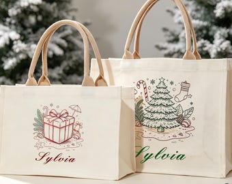 May include: Two off-white canvas tote bags with beige handles. The smaller bag features a red gift box illustration and the name Sylvia in red script. The larger bag displays a Christmas tree, candy cane, and stocking with the name Sylvia in green.