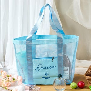 May include: A light blue mesh tote bag with matching handles and a front pocket. The pocket is embroidered with the name "Denise" and a sailboat design. The bag contains a bottle of wine and other items. The bag is on a wooden surface.