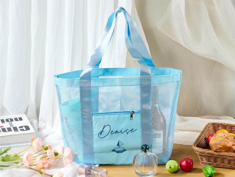 May include: A light blue mesh tote bag with matching handles and a front pocket. The pocket is embroidered with the name "Denise" and a sailboat design. The bag contains a bottle of wine and other items. The bag is on a wooden surface.