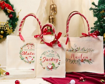 Puede incluir: Tres bolsas de mano de lona color crema con asas y lazos rojos. Cada bolsa presenta un diseño de corona navideña con un nombre en escritura roja. Las bolsas están decoradas con coronas navideñas e ilustraciones de Papá Noel.