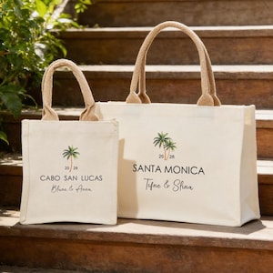 May include: Two cream canvas tote bags with beige handles. The bags feature a palm tree graphic and the text "CABO SAN LUCAS Blune & Anna" and "SANTA MONICA Tifne & Slina". They are placed on wooden steps.