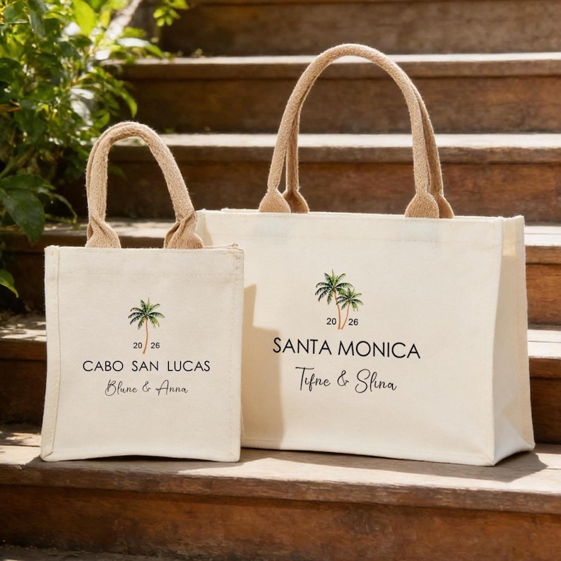 May include: Two cream canvas tote bags with beige handles. The bags feature a palm tree graphic and the text "CABO SAN LUCAS Blune & Anna" and "SANTA MONICA Tifne & Slina". They are placed on wooden steps.