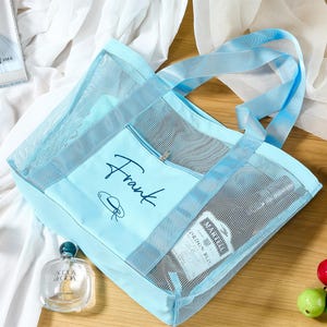 May include: A light blue mesh tote bag with matching handles and trim. The bag has a zippered front pocket with the name "Frank" in script. A bottle of perfume and other items are visible inside.