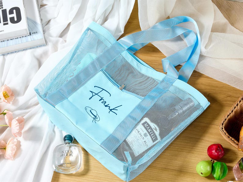 May include: A light blue mesh tote bag with matching handles and trim. The bag has a zippered front pocket with the name "Frank" in script. A bottle of perfume and other items are visible inside.