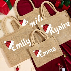 May include: A collection of burlap tote bags with beige handles. Each bag features a white name and a Santa hat graphic in red and white. The bags are arranged on a red surface with Christmas decorations.