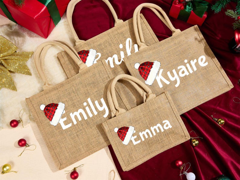May include: A collection of burlap tote bags with beige handles. Each bag features a white name and a Santa hat graphic in red and white. The bags are arranged on a red surface with Christmas decorations.