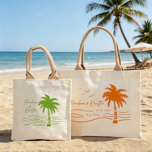 May include: Two off-white tote bags with jute handles are displayed on a sandy beach. The bags feature palm tree graphics and text that reads "Hanhan & Kristen" and "Key West". The larger bag also includes the date "12.10.2026".