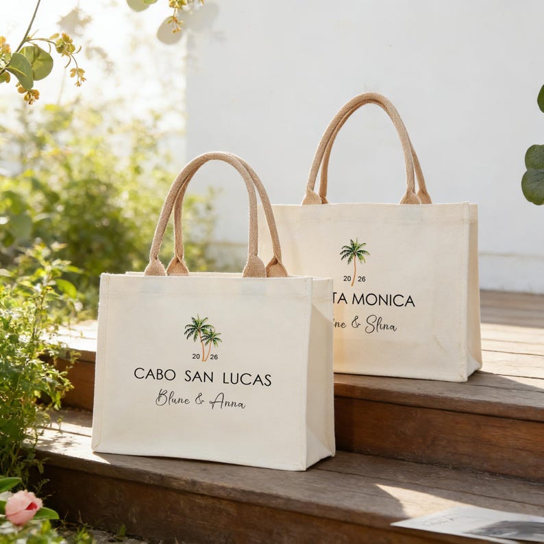 May include: Two cream-coloured canvas tote bags with jute handles. Each bag features a palm tree graphic and text. One bag says "Cabo San Lucas" and the other says "Santa Monica". Both bags have a date and names printed on them.