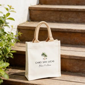 May include: An off-white canvas tote bag with a jute handle. The bag has a palm tree graphic and the text "CABO SAN LUCAS" with the names "Blane & Anna" below. The bag is placed on wooden steps.